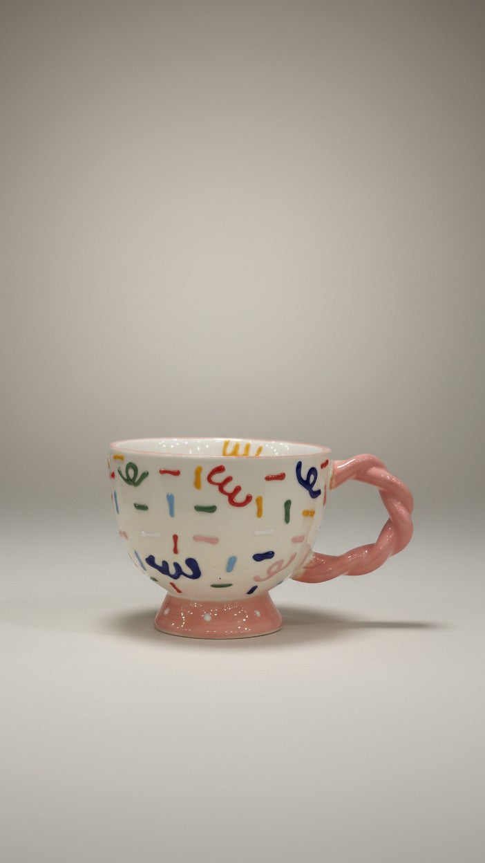 Asoula white ceramic mug with colorful confetti pattern and pink twisted handle. Perfect for coffee or tea.