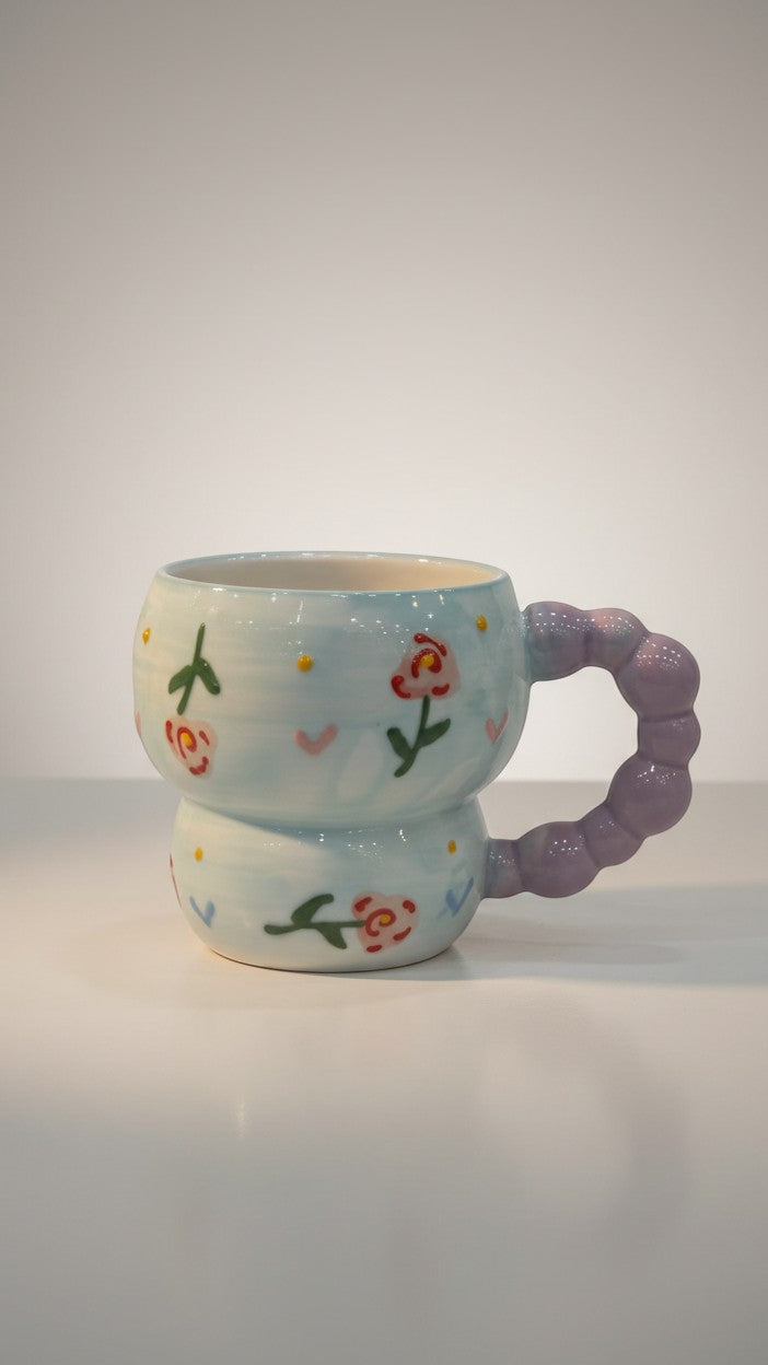 Asoula light blue ceramic mug with a whimsical floral design and a purple bubble handle. Perfect for coffee or tea.