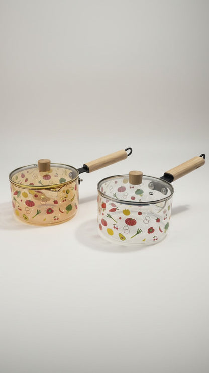 Asoula clear glass saucepan with lid, featuring a playful vegetable print and wooden handle. Oven-safe for versatile cooking.