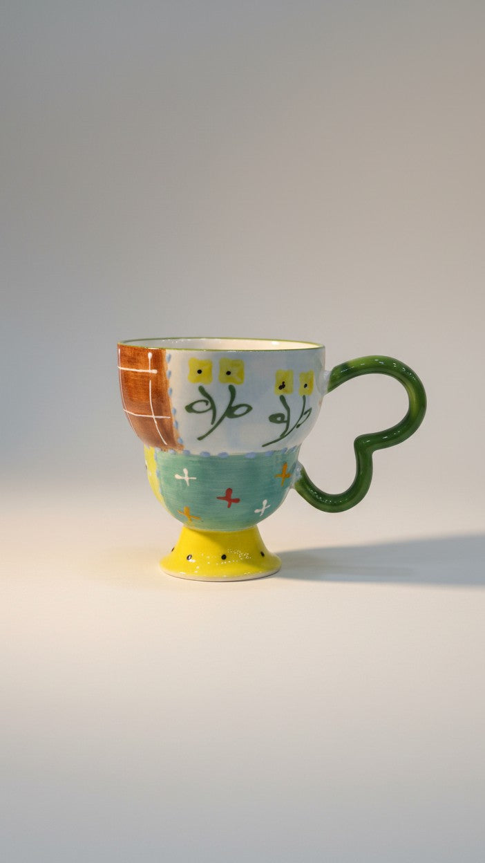 Asoula ceramic mug with a unique patchwork design featuring floral motifs, hearts, and crosses in yellow, green, and red.