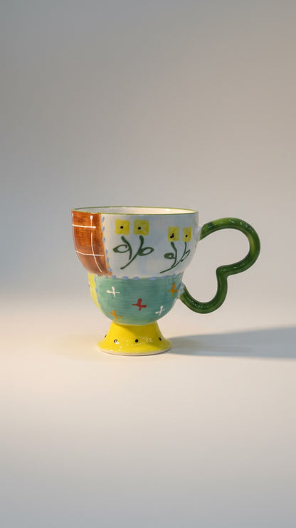 Asoula ceramic mug with a unique patchwork design featuring floral motifs, hearts, and crosses in yellow, green, and red.