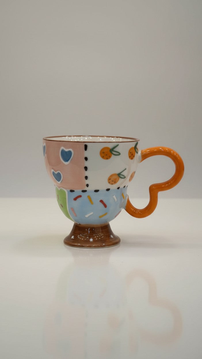 Asoula ceramic mug with patchwork design featuring hearts, oranges, and sprinkles. Features a unique orange loop handle.