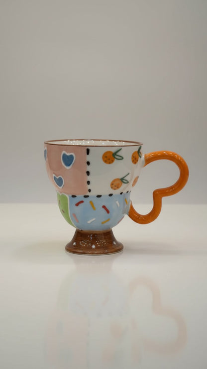 Asoula ceramic mug with patchwork design featuring hearts, oranges, and sprinkles. Features a unique orange loop handle.
